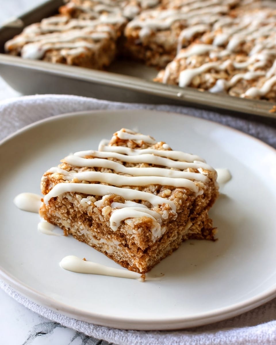 A square piece of baked oat bar sits on a smooth white plate, showing three visible layers: a rough, golden-brown oat crust on top with a crunchy texture, a dense, slightly lighter brown oat middle layer with a chewy appearance, and a moist bottom layer that blends slightly with the middle. The top layer is drizzled with a creamy white icing in uneven lines, adding a soft contrast to the textured surface. In the background, part of a larger tray filled with similar oat bars, also drizzled with white icing, is visible resting on a cloth over a white marbled surface. photo taken with an iphone --ar 4:5 --v 7