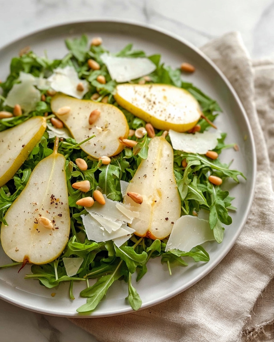 A white plate holds a fresh salad with a base layer of dark green arugula leaves spread evenly. On top, several light yellow-green pear slices with brown speckled skin are arranged in a scattered pattern. Small golden pine nuts are sprinkled across the salad, adding texture and contrast. Thin, off-white shavings of cheese rest gently over the pears, and a light sprinkling of black pepper dots the entire dish. The plate is set on a soft beige cloth on a white marbled surface. photo taken with an iphone --ar 4:5 --v 7