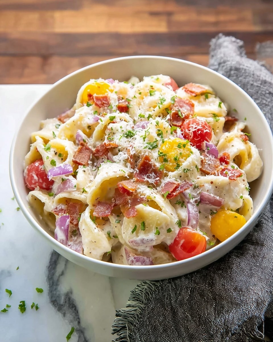 A white bowl filled with creamy pasta salad sits on a white marbled surface, showing layers of folded pasta coated in a white sauce. Scattered throughout are round slices of red and yellow cherry tomatoes and small pieces of crispy reddish-brown bacon. Thin slices of purple-red onion add texture, and the dish is topped with grated white cheese and small sprinkles of green herbs. A gray cloth napkin with frayed edges is placed to the side of the bowl. Photo taken with an iphone --ar 4:5 --v 7