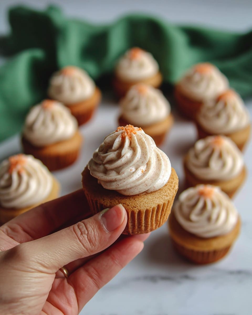 The image shows a close-up of a small cupcake held by a woman's hand, with a total of two layers: a light brown cake base and a thick swirl of beige cream with fine specks on top. In the background, about a dozen more cupcakes are arranged closely together, each with the same two layers and topped with a small orange sprinkle. The scene is set on a white marbled surface with a green cloth softly blurred in the background, creating a cozy and inviting feel. Photo taken with an iphone --ar 4:5 --v 7
