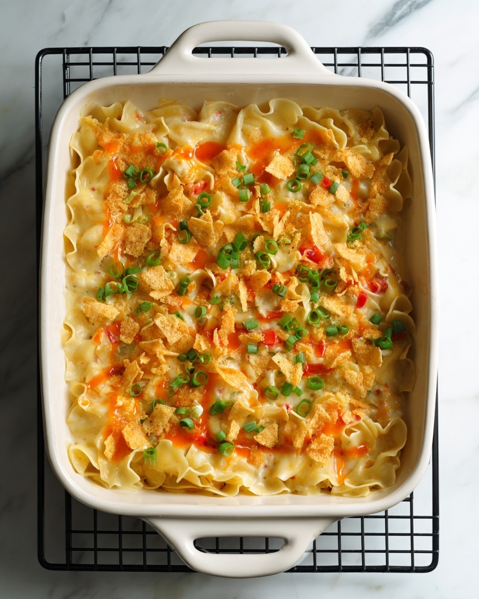 A baked dish in a white rectangular casserole with handles, set on a black cooling rack on a white marbled surface, shows three clear layers: the bottom layer has a pale yellow, soft egg noodle base with ruffled edges; the middle layer contains white, creamy sauce mixed with pieces of red bell pepper and light-colored vegetables; the top layer is sprinkled with broken, golden yellow crispy chips and chopped green onions, with a drizzle of an orange sauce adding a glossy shine across the top. photo taken with an iphone --ar 4:5 --v 7