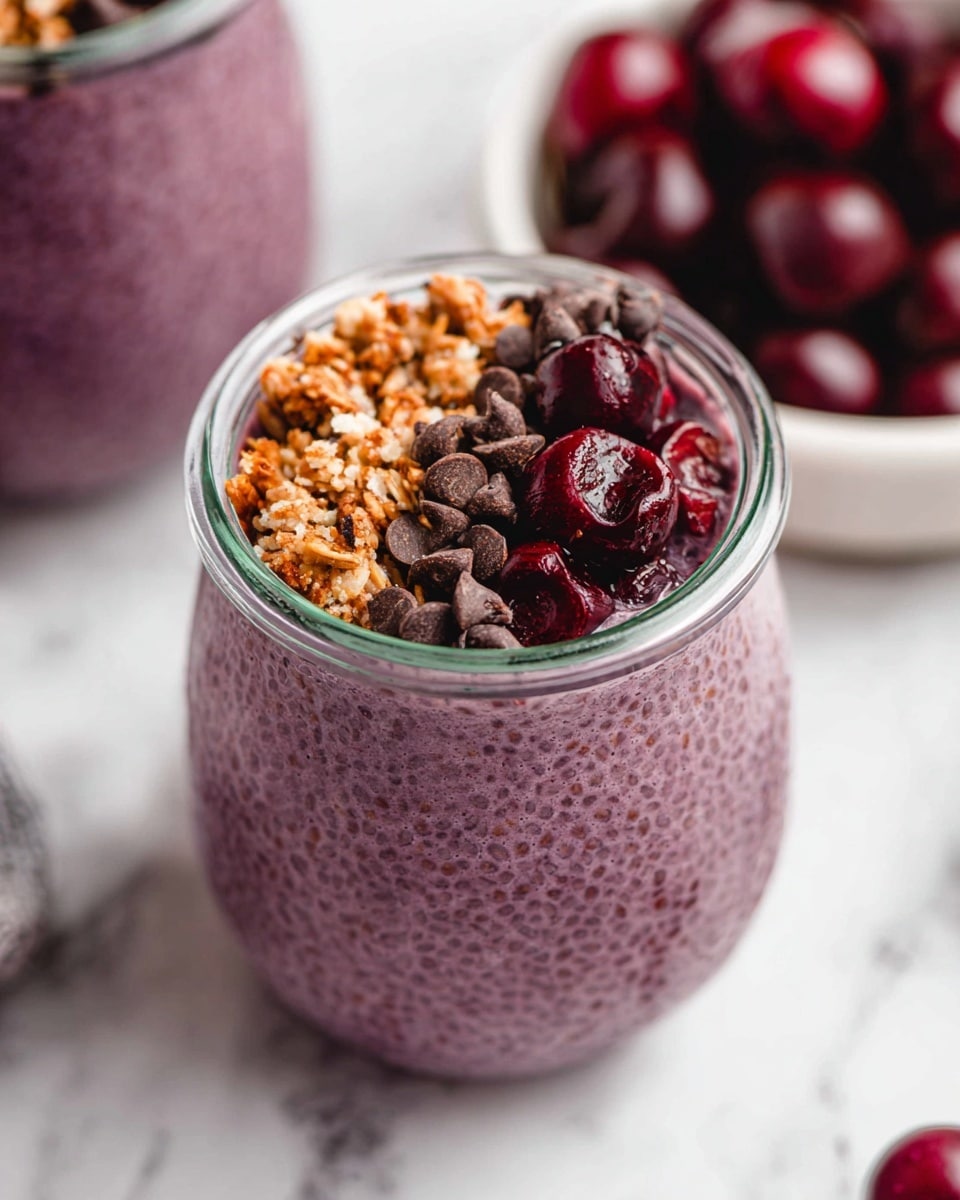 A glass jar filled with a purple chia seed pudding, showing its smooth and slightly bumpy texture from the soaked chia seeds, topped with three toppings in sections: light brown granola with a crunchy texture, dark brown chocolate chips, and dark red chopped cherries with a juicy look, all arranged neatly on top. In the background, there is a blurred glass jar with more purple chia pudding and a white bowl filled with whole dark red cherries, placed on a white marbled surface. Photo taken with an iphone --ar 4:5 --v 7
