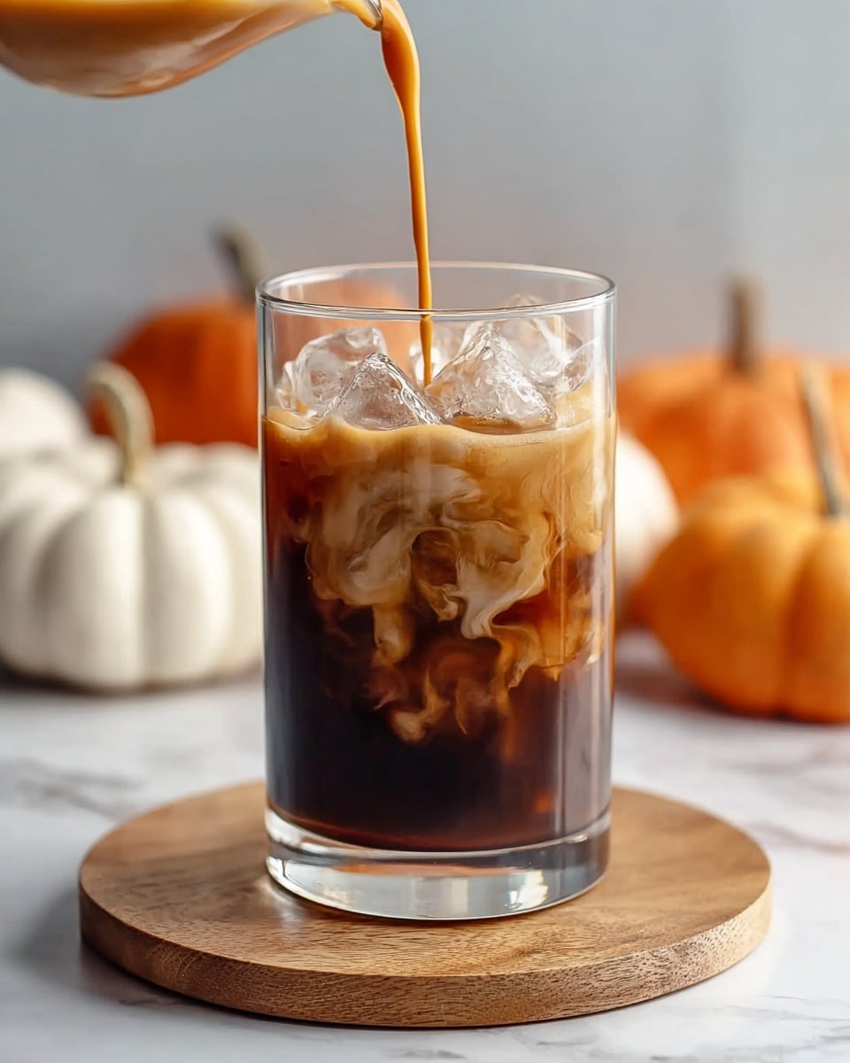 A clear glass filled with dark brown iced coffee and large ice cubes near the top shows a layer of light brown creamy foam forming as a golden iced coffee liquid is poured into it from above. The glass sits centered on a round wooden board set on a white marbled surface, with blurred white and orange pumpkins in the soft-focused background. Photo taken with an iphone --ar 4:5 --v 7