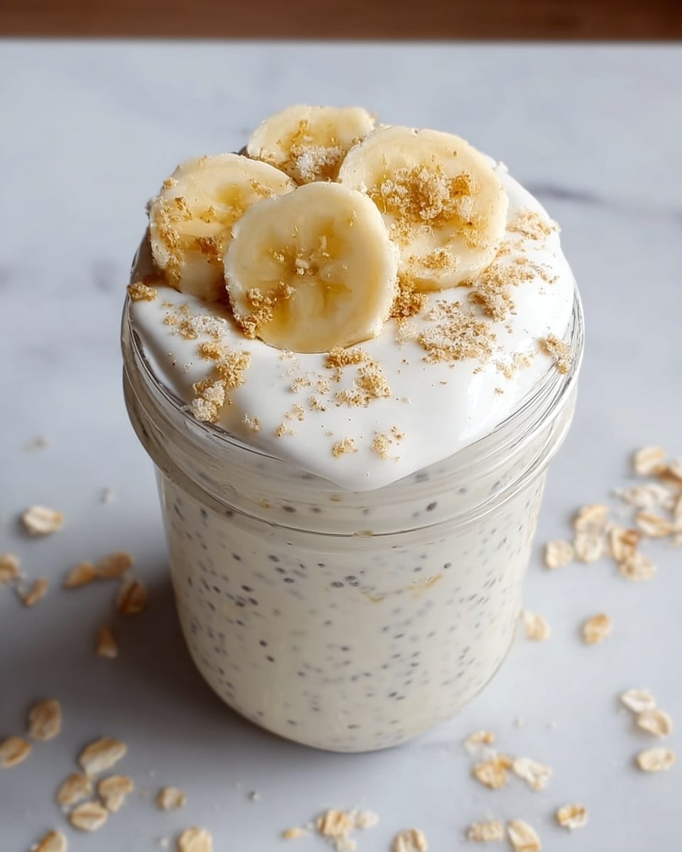 A clear glass jar filled with a creamy mixture that has tiny black chia seeds evenly spread throughout. The top layer is a thick, white cream that almost overflows at the edges. On top of the cream, there are five banana slices placed close together, with light brown crumbs sprinkled over the banana and cream. The jar sits on a white marbled surface with scattered oat flakes nearby. Photo taken with an iphone --ar 4:5 --v 7