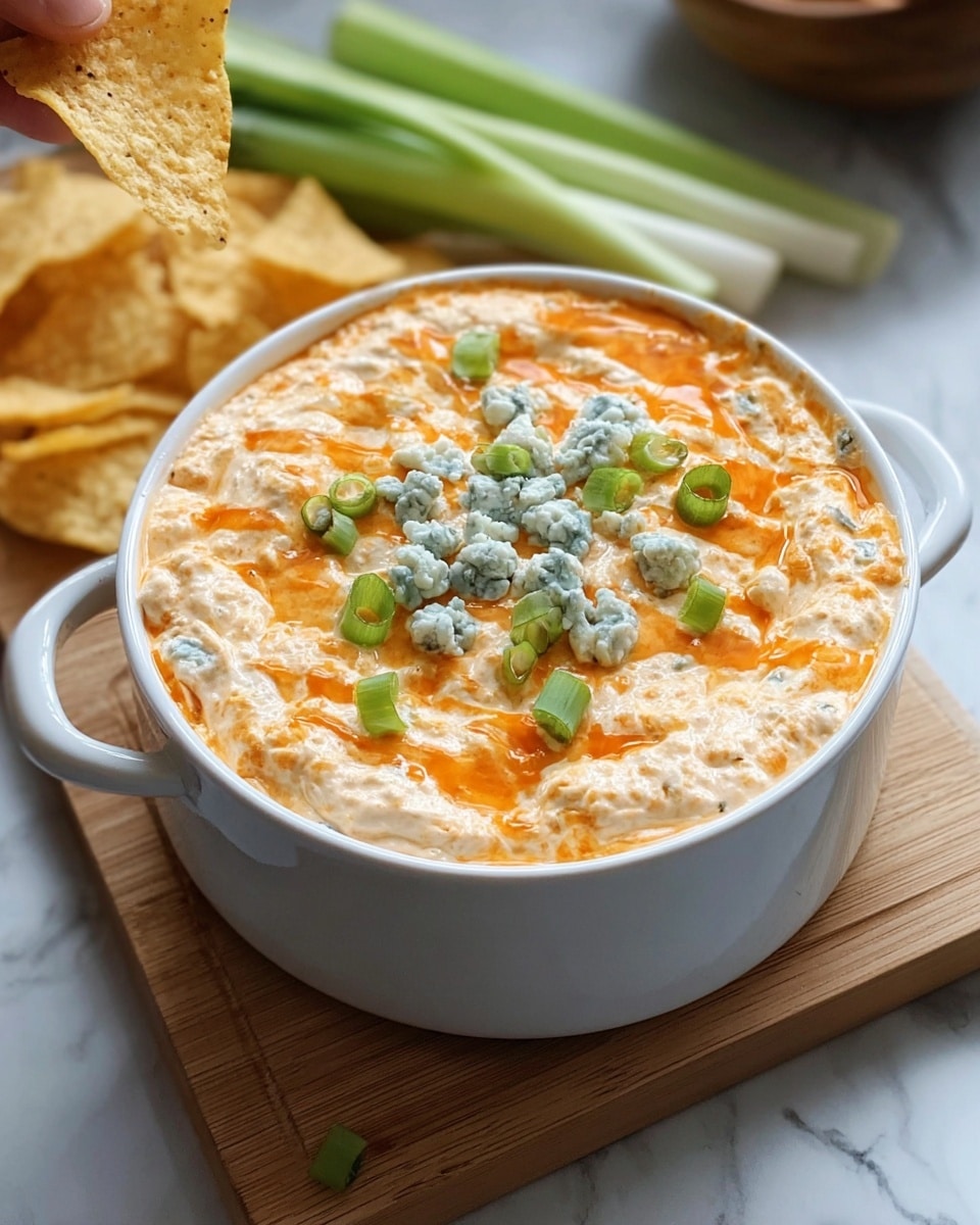A white bowl with a handle holds a creamy dip with two layers, the bottom layer being a thick, pale orange cheese mix with a slightly lumpy texture, and the top layer thinner but still rich, drizzled with a brighter orange sauce. The dip is topped with scattered finely chopped green onions and small dollops of crumbly blue cheese in the center. The bowl sits on a light wooden board, with green onions and tortilla chips resting in the background on a white marbled surface. A woman's hand holding a chip is partially visible at the top left corner. photo taken with an iphone --ar 4:5 --v 7