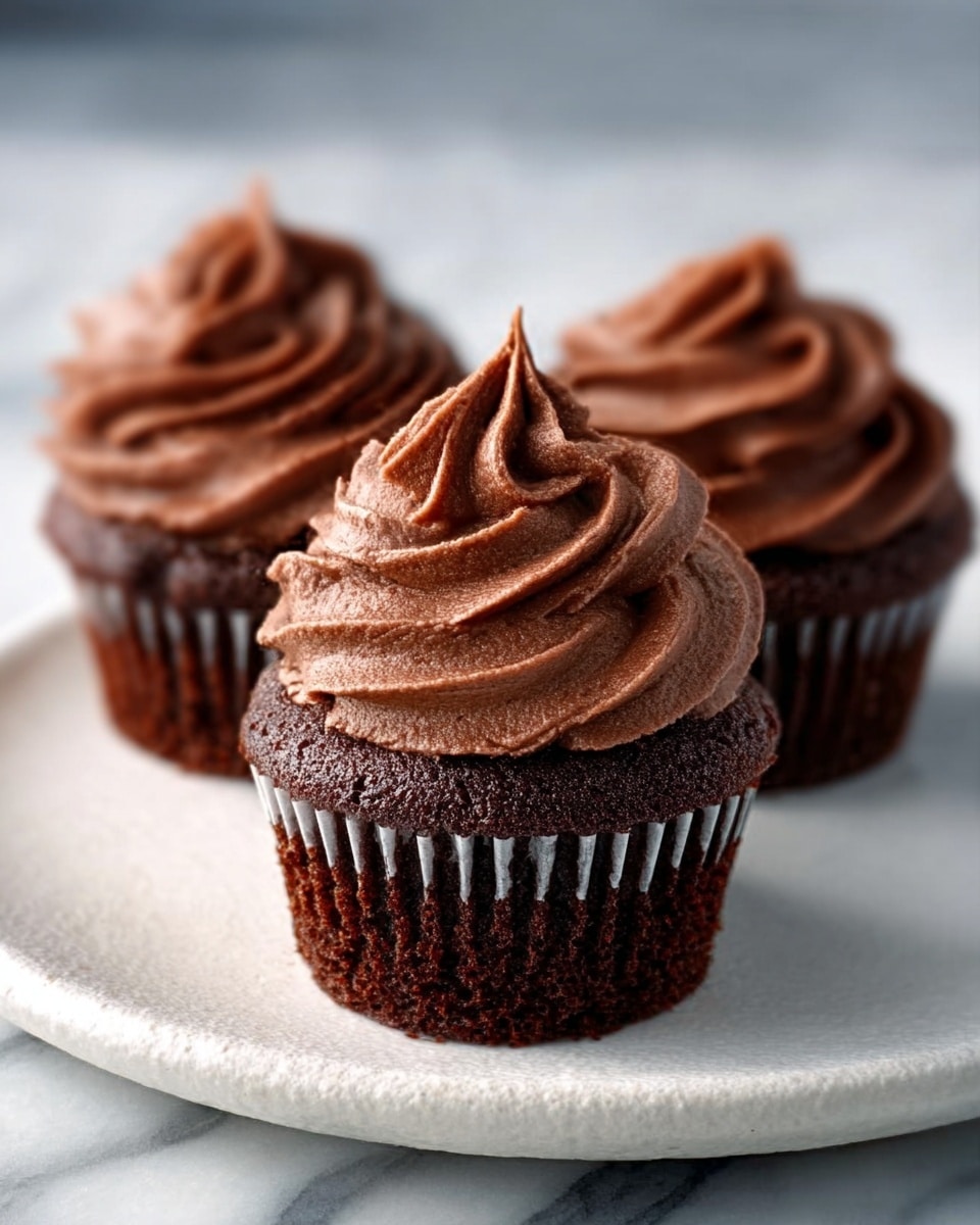 The image shows a close-up of a chocolate cupcake with three visible layers: the bottom dark brown chocolate cake layer in a ridged brown paper liner, topped by a thick layer of smooth, light brown chocolate frosting that forms a swirl with soft peaks, and sprinkled with small, dark brown chocolate sprinkles scattered evenly on the frosting. Behind the main cupcake, there are several more chocolate cupcakes with similar frosting swirls, all sitting on a white plate with a black edge, placed against a white marbled textured surface. photo taken with an iphone --ar 4:5 --v 7