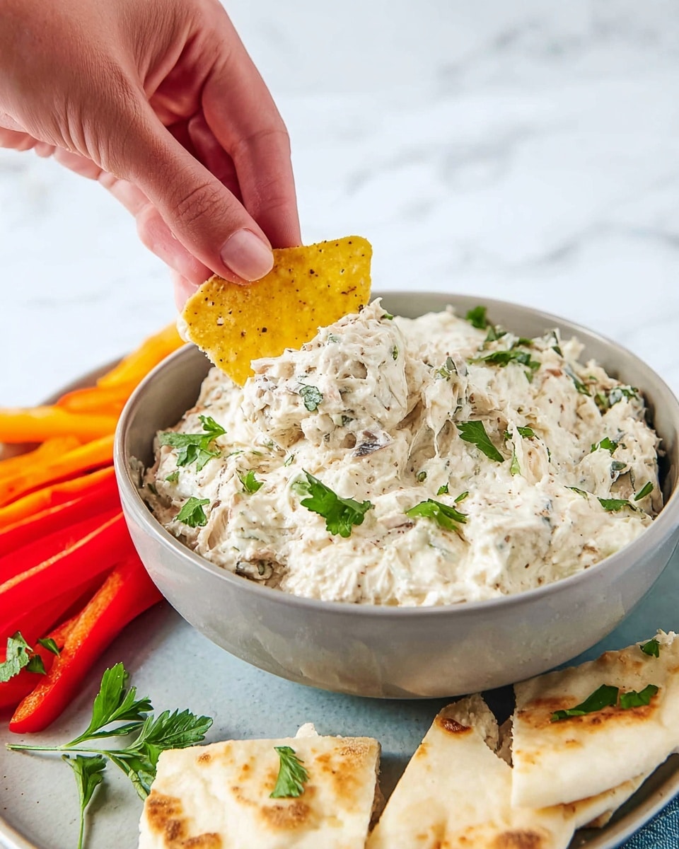 A light gray bowl holds a creamy, chunky dip with a thick texture, speckled with small bits of herbs and other ingredients, topped with fresh green parsley pieces scattered on the surface. A yellow tortilla chip is scooping the dip, held by a woman's hand above the bowl. Around the bowl on a white plate, there are cut pieces of soft white pita bread garnished with parsley, and colorful red and orange bell pepper strips are partially visible on the left side. The setting is on a white marbled surface. photo taken with an iphone --ar 4:5 --v 7