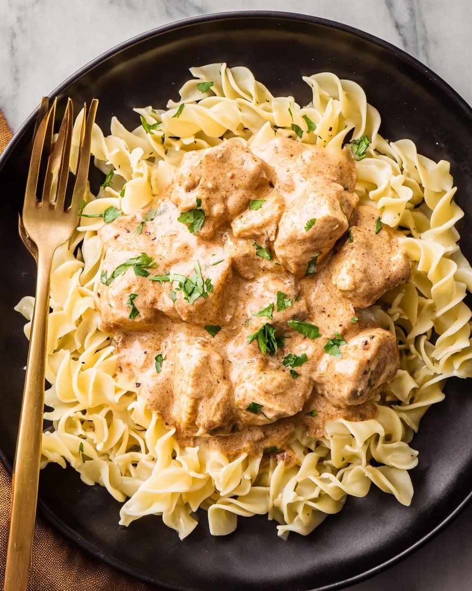A round black plate holds a bed of pale yellow egg noodles with a slightly twisted shape, arranged loosely around the edges. On top, there are several chunks of tender chicken covered in a creamy, light brown sauce with specks of seasoning, giving it a smooth and rich texture. Small green parsley leaves are sprinkled over the sauce for a fresh burst of color. A gold fork is placed on the left side, resting on the edge of the plate. The whole scene is set against a white marbled surface. photo taken with an iphone --ar 4:5 --v 7
