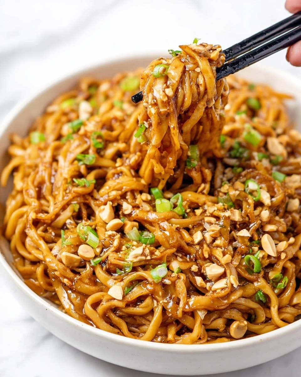 The image shows a white bowl filled with noodles coated in a brown sauce, giving them a glossy, textured look. The noodles are thick and slightly curly, mixed well with small pieces of crushed nuts and chopped green onions scattered evenly on top. A pair of black chopsticks, held by a woman's hand, lifts a tangled bundle of noodles from the bowl, showcasing the sauce and toppings grasped within. The background has a white marbled texture that enhances the warm colors of the food. photo taken with an iphone --ar 4:5 --v 7