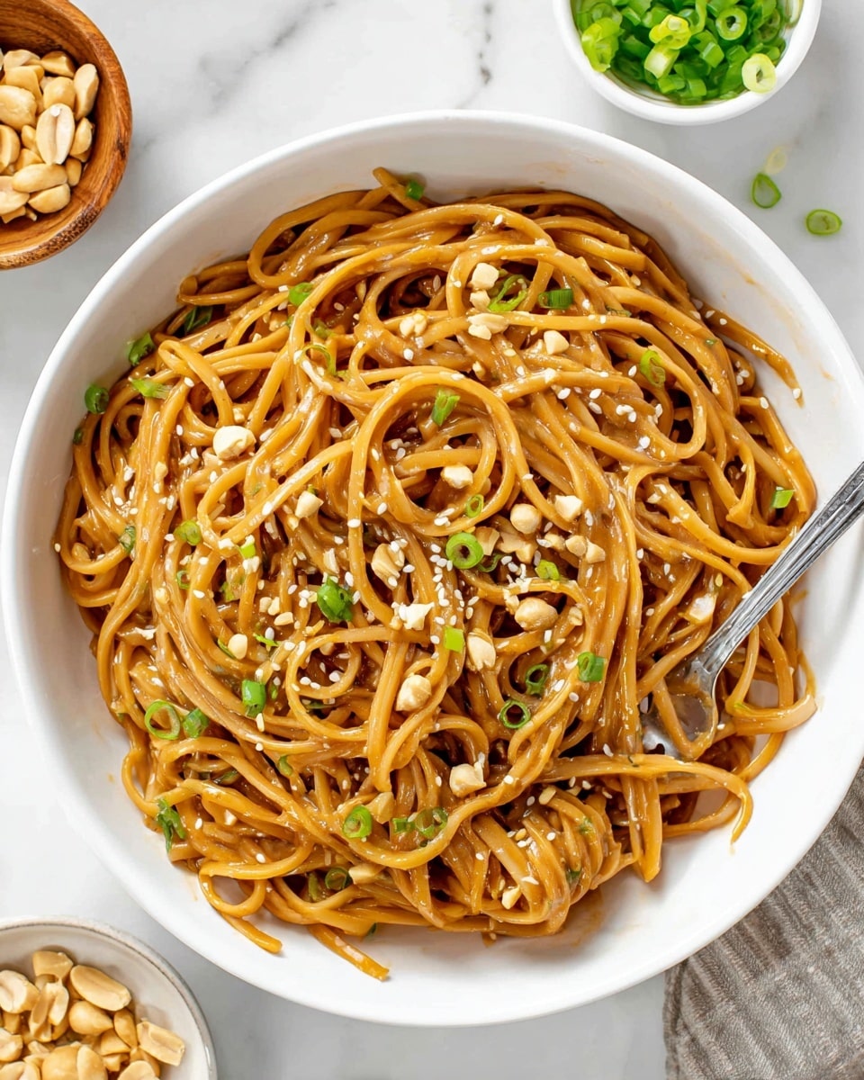 A white bowl filled with a single layer of glossy, light brown noodles coated in a thick, shiny peanut sauce. The noodles are long and slightly twisted, mixed evenly with small green onion pieces scattered on top. Light tan crushed peanuts and white sesame seeds are sprinkled throughout the dish, adding small pops of texture and color. A silver fork rests inside the bowl, partially covered by the noodles. The bowl is placed on a white marbled surface with a small white bowl of green onion slices and a small wooden bowl filled with chopped peanuts nearby. Photo taken with an iphone --ar 4:5 --v 7