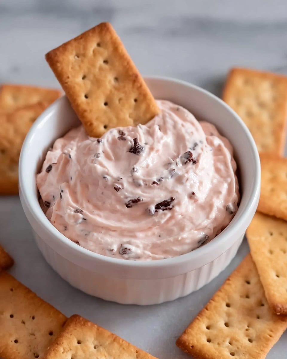 A white small bowl filled with a creamy pink dip mixed with small dark bits, likely chocolate chips, with a smooth, slightly swirled texture on top. One rectangular golden-brown cracker is placed upright inside the dip, partially submerged and showing small holes. Around the bowl, there are several more of the same golden-brown rectangular crackers scattered on a white marbled surface. Photo taken with an iphone --ar 4:5 --v 7