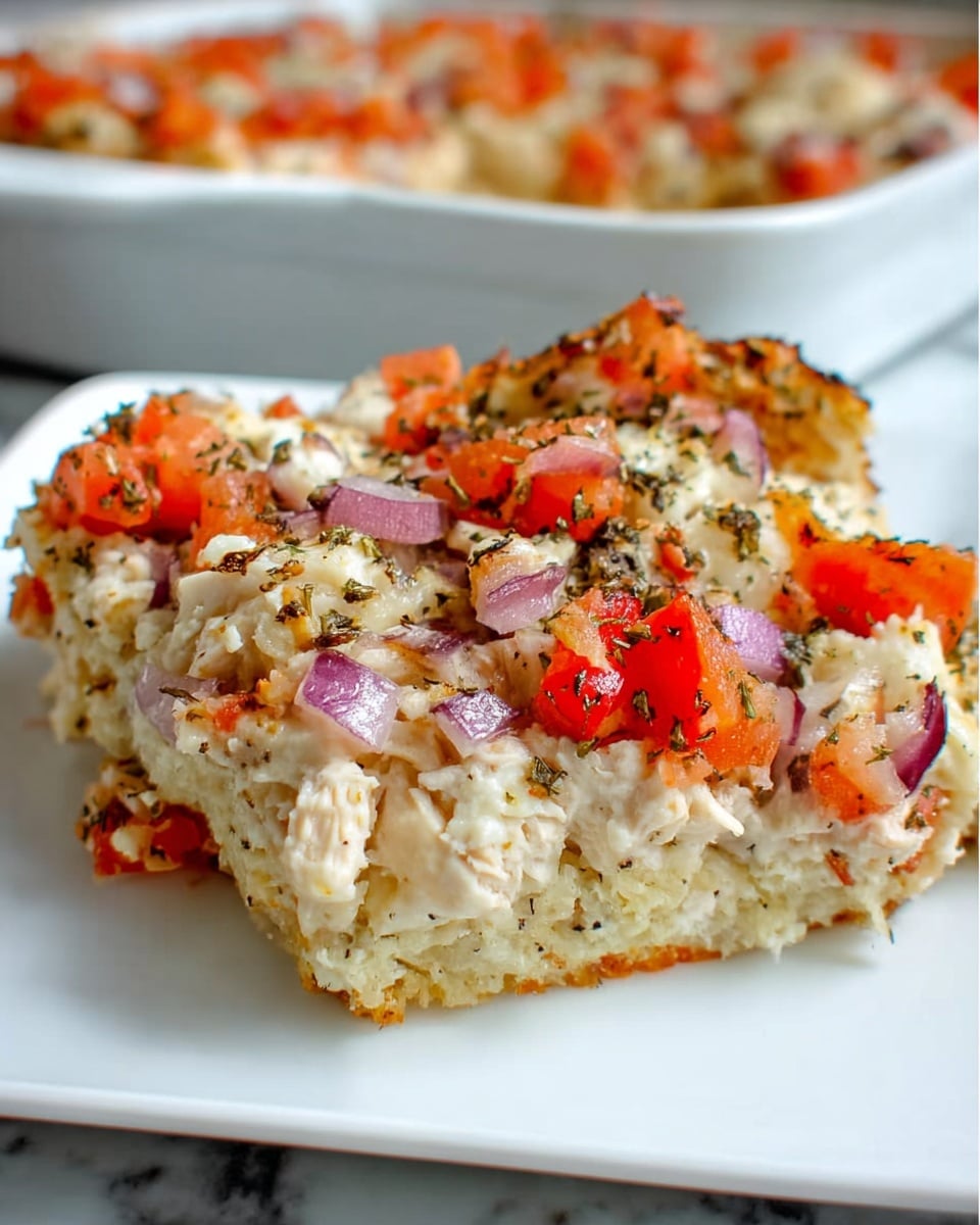 A close-up of a single serving of a baked casserole on a square white plate, showing a thick bottom layer made of shredded light beige cooked chicken, topped with a creamy, slightly crumbly white cheese mixture spread evenly. On top, there are diced bright red tomatoes and thin purple onion slices scattered throughout, all sprinkled with small greenish herb flakes and black pepper. The texture looks soft and chunky with slightly browned edges. The background is a blurred casserole dish full of more servings, all set on a white marbled surface. Photo taken with an iphone --ar 4:5 --v 7
