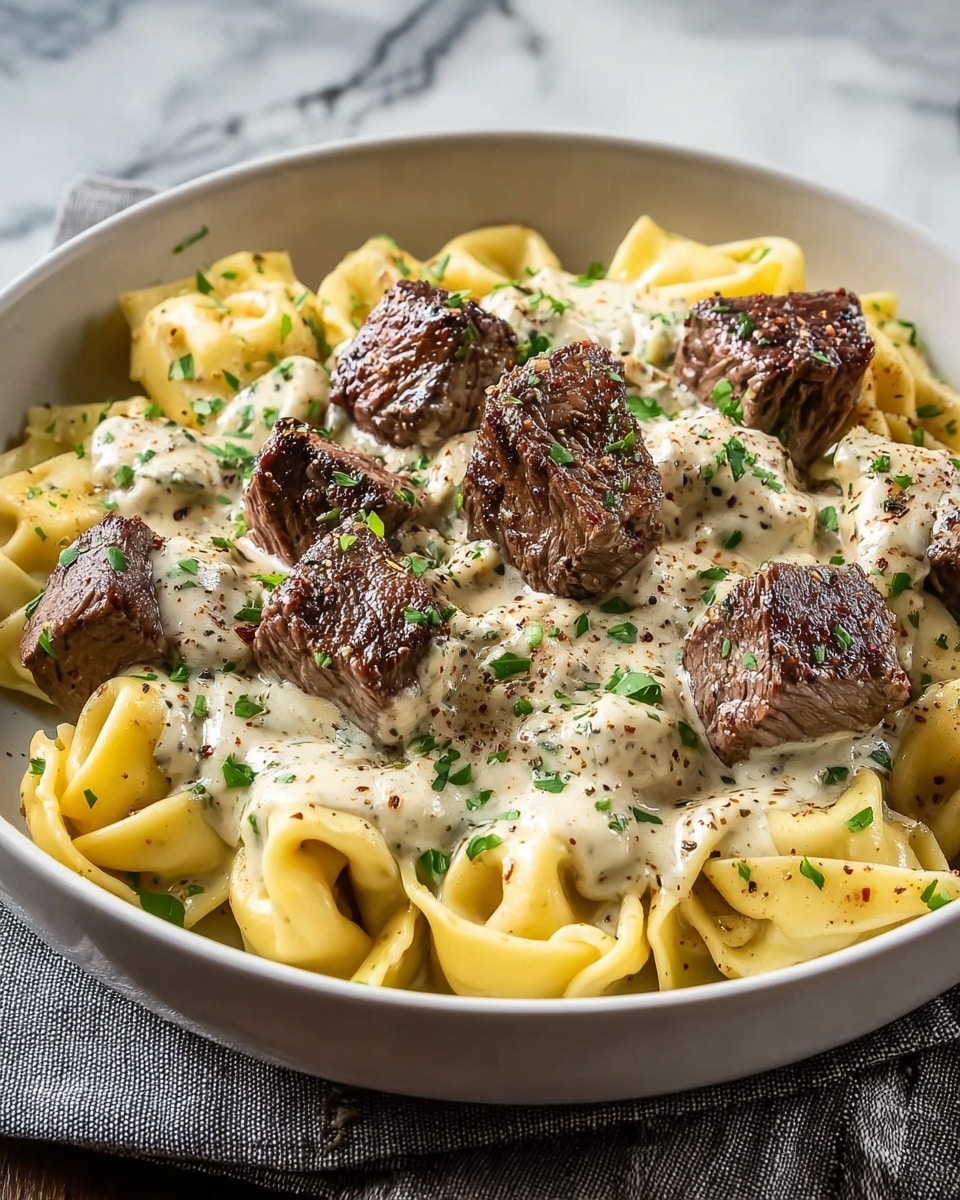 A white bowl filled with tortellini pasta covered in a smooth, creamy white sauce, topped by several chunks of browned, grilled steak with a slightly charred texture. The tortellini are light yellow with a soft, folded shape, and the sauce has a rich, glossy look sprinkled with cracked black pepper and small pieces of fresh green herbs scattered on all layers. The bowl sits on a gray cloth on a white marbled surface. photo taken with an iphone --ar 4:5 --v 7