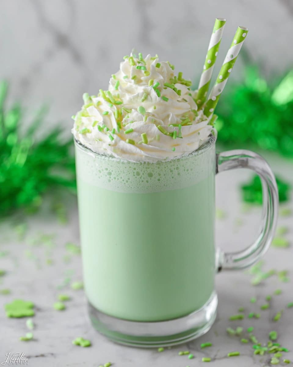 A clear glass mug filled with a smooth, light green drink takes center focus. The drink fills most of the mug, topped with a thick layer of white whipped cream that is swirled in soft peaks, adorned with small round and rod-shaped green sprinkles. Two green straws with white polka dots stick out from the whipped cream on a white marbled surface background, with faint green decorations blurred in the back. photo taken with an iphone --ar 4:5 --v 7