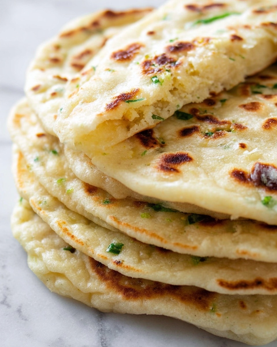 A close-up view of a stack of five round flatbreads with a soft, slightly puffy texture and golden brown spots on the surface. The top flatbread is slightly folded, showing its airy inside with a light yellow color. Visible inside the flatbreads are small bits of green herbs, mostly near the edges. The flatbreads have a glistening shine, suggesting they are lightly brushed with butter or oil. The stack is placed on a white marbled surface. Photo taken with an iphone --ar 4:5 --v 7