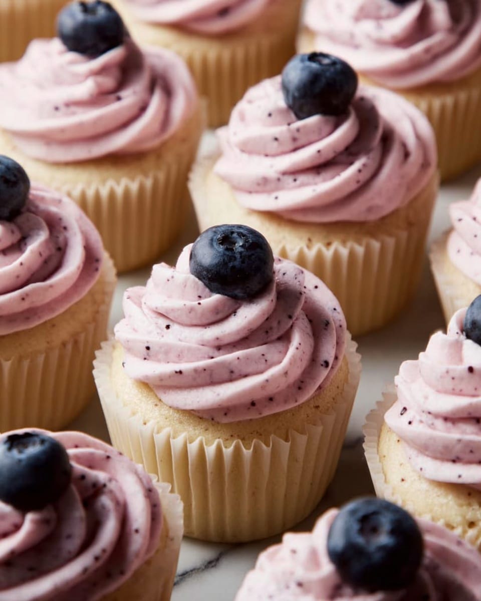 A close-up view shows many vanilla cupcakes in white paper cups, each topped with a swirl of light pink frosting mixed with small dark specks, creating a creamy texture. On top of the frosting, a single fresh dark blue blueberry is placed, adding a touch of color contrast. The cupcakes sit closely together on a white marbled surface, filling the entire frame with soft natural light highlighting the creamy frosting's smooth peaks and the blueberries’ glossy surface. Photo taken with an iphone --ar 4:5 --v 7