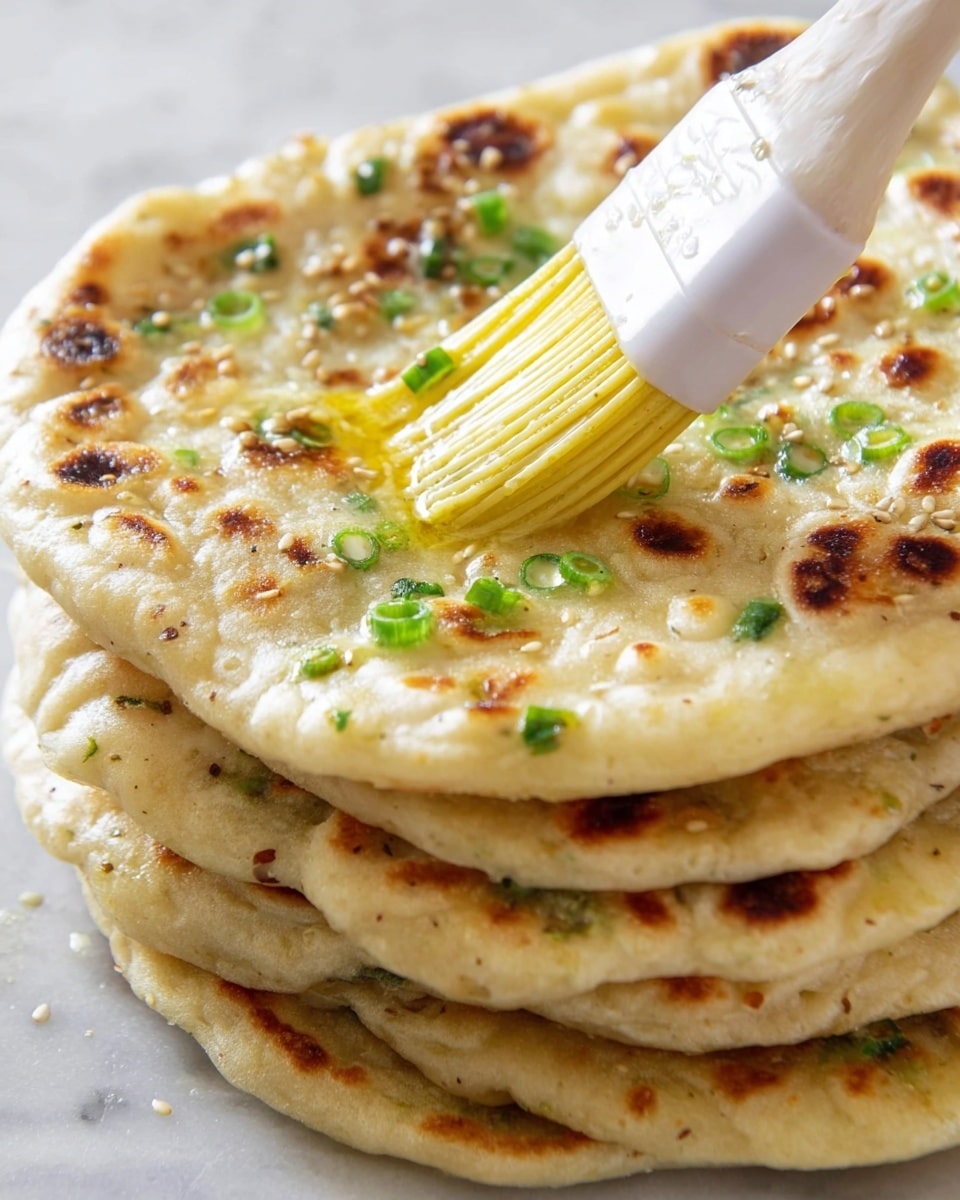 A stack of five flatbreads with light golden and slightly browned spots, each layer thin and soft, with chopped green onions sprinkled on top and embedded in the bread. The top flatbread is being brushed with yellow melted butter using a white pastry brush, adding a shiny texture. The flatbreads show a slightly uneven surface with some bubbles and cooked seeds, and the setting is on a white marbled texture. photo taken with an iphone --ar 4:5 --v 7