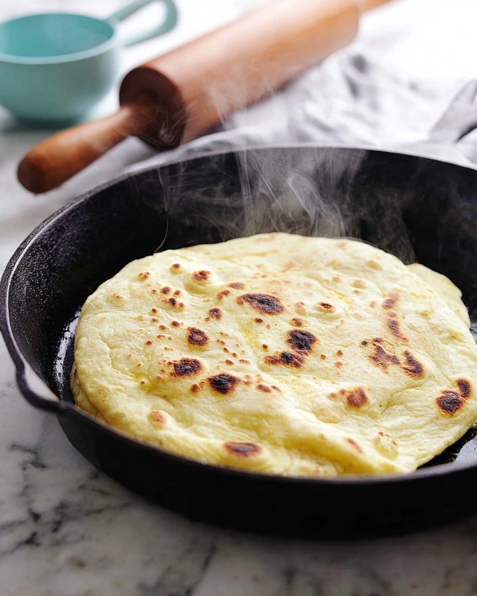 A single thick flatbread with a pale yellow color and a soft, slightly puffed texture is cooking in a black cast iron pan. It has unevenly scattered brown char spots on the surface, showing it is freshly toasted. Wisps of steam rise from the flatbread, suggesting it is hot and fresh. The pan sits on a white marbled surface with a light blue measuring cup and a wooden rolling pin blurred in the background, adding a cozy kitchen feel. photo taken with an iphone --ar 4:5 --v 7