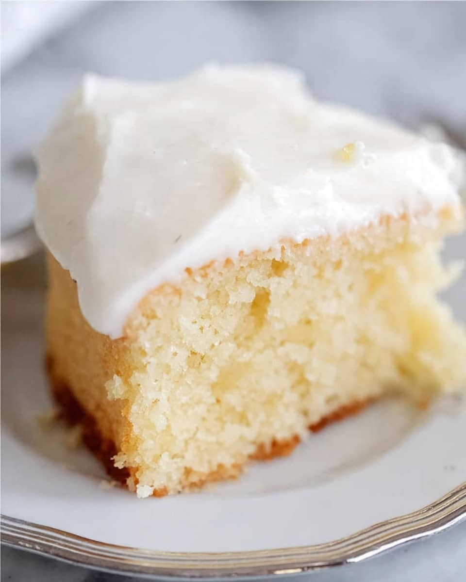 A close-up of a single slice of light yellow cake with a soft, moist texture, sitting on a white plate with a silver rim. The cake has one thick layer topped with a smooth, white frosting layer that looks creamy and slightly fluffy, covering the top evenly and spilling slightly on the sides. The bottom edge of the cake shows a slightly browned crust. The background is a white marbled texture. photo taken with an iphone --ar 4:5 --v 7