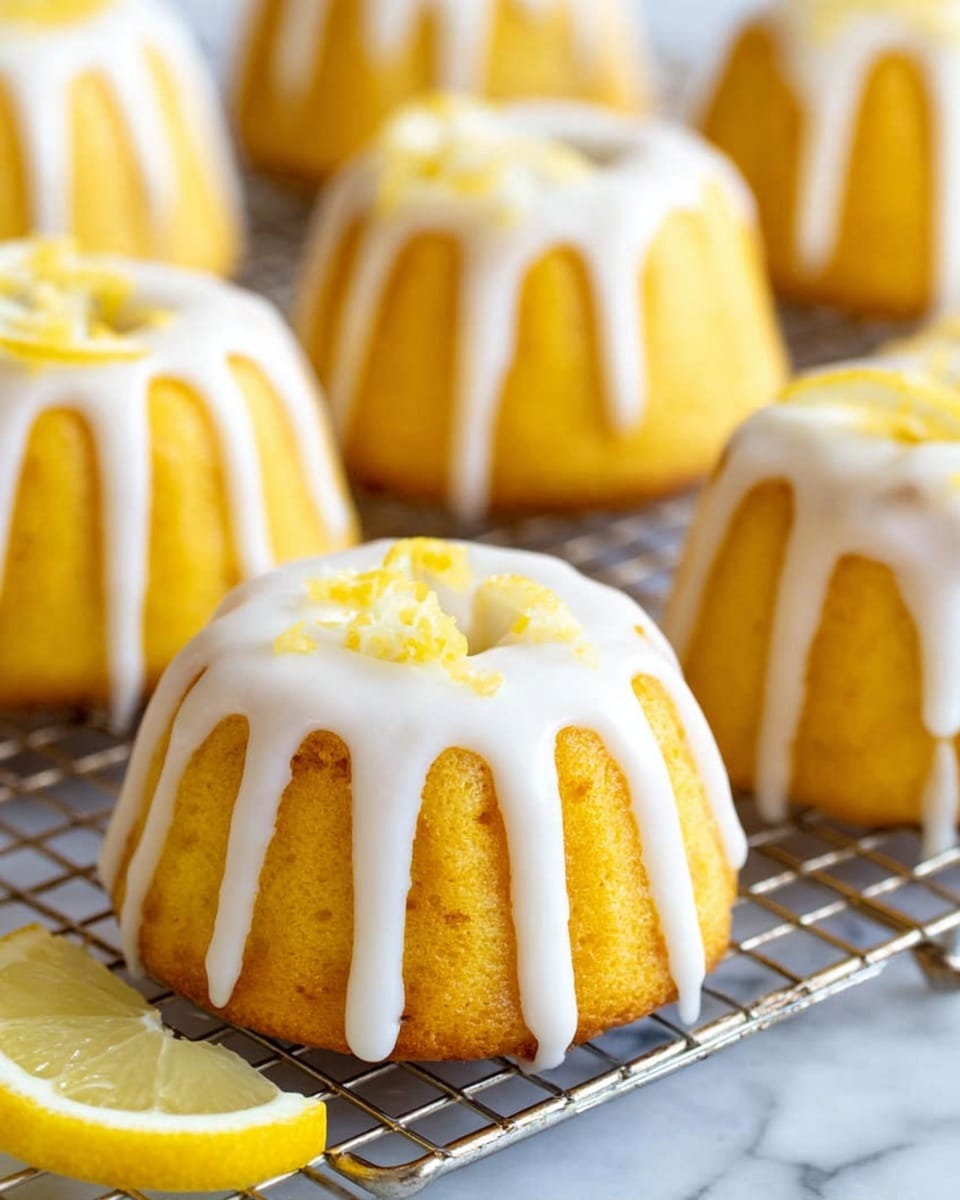 The image shows several small round bundt cakes with a bright yellow color, each topped with thick white icing dripping evenly down the sides in stripes. The cakes sit on a silver cooling rack, and there is a small wedge of lemon resting beside one of the cakes, adding a fresh touch. The yellow cakes have a smooth texture, and the icing is shiny and creamy. The background is a white marbled surface. photo taken with an iphone --ar 4:5 --v 7