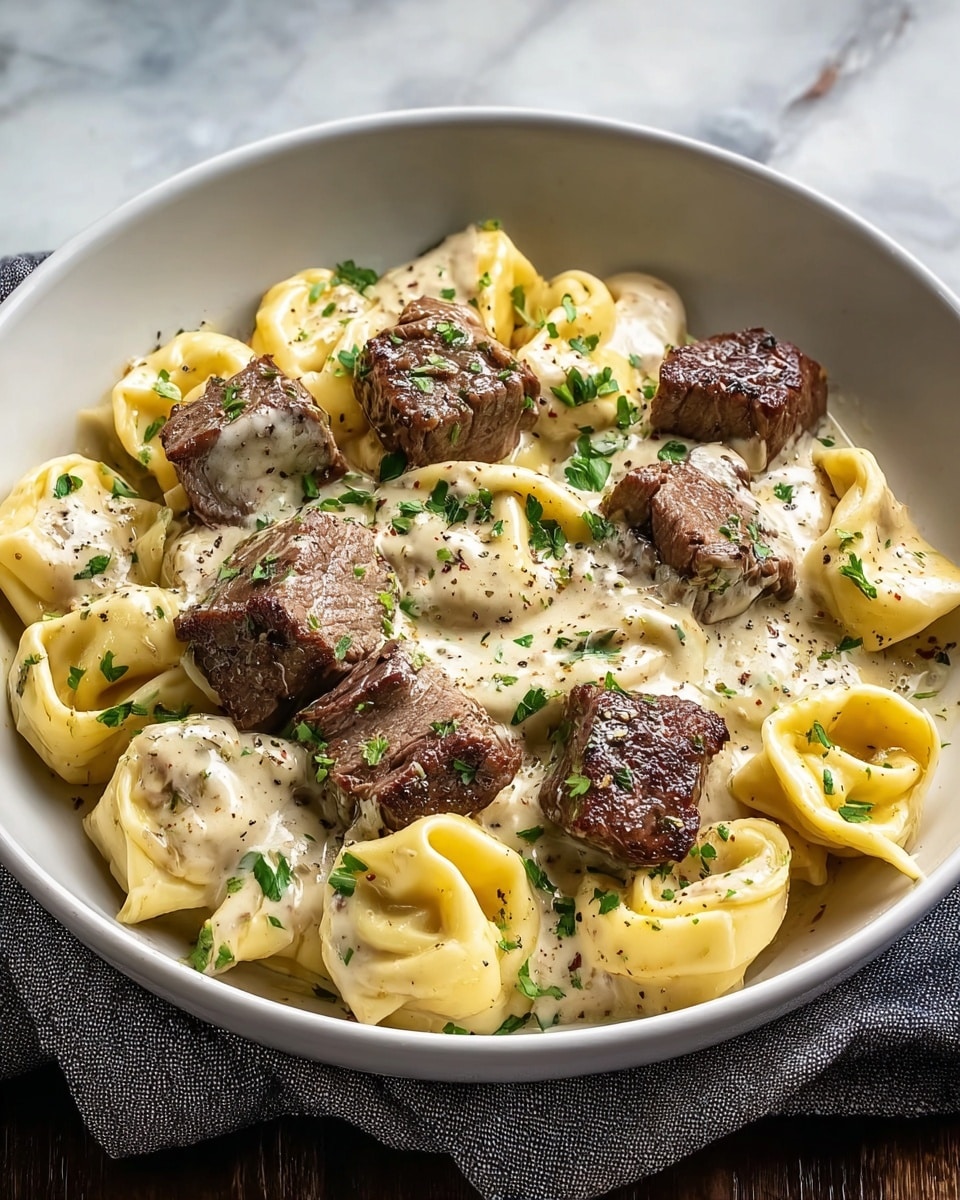 A white bowl holds a dish made of three layers: the bottom layer is light yellow tortellini pasta with smooth, folded shapes; on top is a creamy white sauce speckled with black pepper; the top layer shows browned chunks of grilled steak with a slightly crispy texture and a rich brown color. The dish is finished with small pieces of chopped green herbs scattered over everything. The bowl sits on a gray cloth beneath it with a white marbled texture in the background photo taken with an iphone --ar 4:5 --v 7