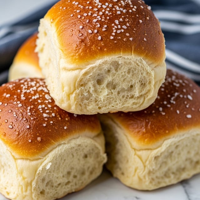 The image shows three golden brown bread rolls stacked close together, with the top roll prominently in focus. Each roll has a soft, fluffy texture with a slightly shiny crust sprinkled with coarse salt crystals. The bread has a light yellowish inside visible at the edges, showing a tender and airy crumb. The background includes a subtle hint of a dark blue and white striped cloth, with the whole scene resting on a white marbled texture. photo taken with an iphone --ar 4:5 --v 7