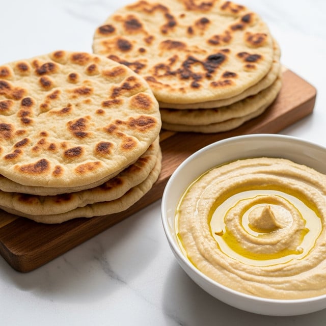 A stack of flat, round, golden-brown flatbreads with uneven, rustic spots of darker brown on their surface sits on a wooden board, showing soft texture and slight puffiness. Next to the flatbreads is a white bowl filled with creamy beige hummus, glossy with a swirl on top and a small pool of olive oil. The scene is set on a white marbled surface that adds a clean, elegant background to the warm colors of the food. photo taken with an iphone --ar 4:5 --v 7