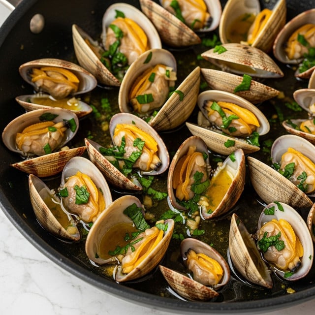 A close-up view of a black pan filled with cooked clams, each clam shell opened showing soft, orange and beige clam meat inside, covered with fresh green chopped herbs. The clams sit in a glossy, buttery sauce with visible bits of garlic and herbs, creating a rich texture. The shells have a mix of light beige and darker brown colors, some with a slightly charred look, and the sauce pools at the bottom of the pan. The pan is placed on a white marbled surface. photo taken with an iphone --ar 4:5 --v 7