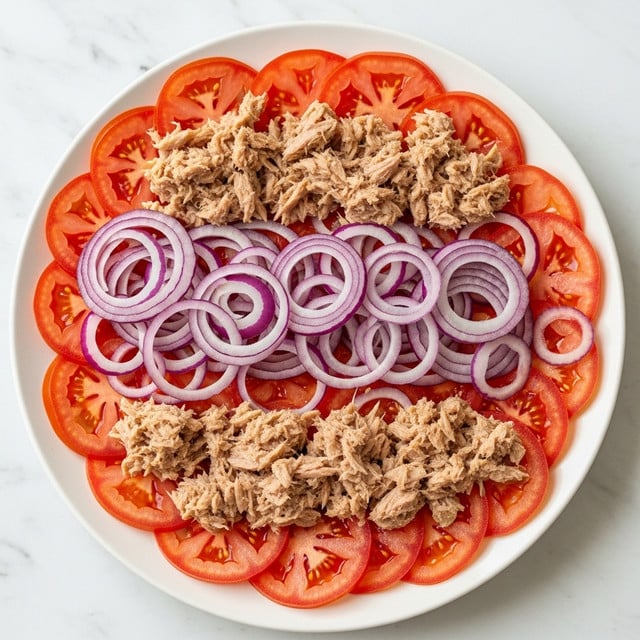 The image shows a white plate with a fresh salad arranged in three visible layers. The bottom layer is made of thin, round red tomato slices spread evenly across the plate. On top of the tomatoes, there is a layer of red onion rings that are spread out in a slightly overlapping way. The top layer consists of small clumps of light brown tuna scattered all over the onions and tomatoes. The background is a white marbled surface. Photo taken with an iphone --ar 4:5 --v 7