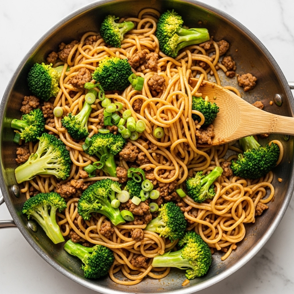 A metal pan filled with a mixed dish of light brown noodles, bright green broccoli florets, and small pieces of cooked ground meat spread evenly throughout. The noodles have a smooth texture and are coated lightly with sauce, while the broccoli is vibrant and firm. Chopped green onions sprinkled on top add a fresh touch, and a wooden spatula is lifting a portion of the noodles and broccoli from the pan. The pan sits on a white marbled surface. photo taken with an iphone --ar 4:5 --v 7