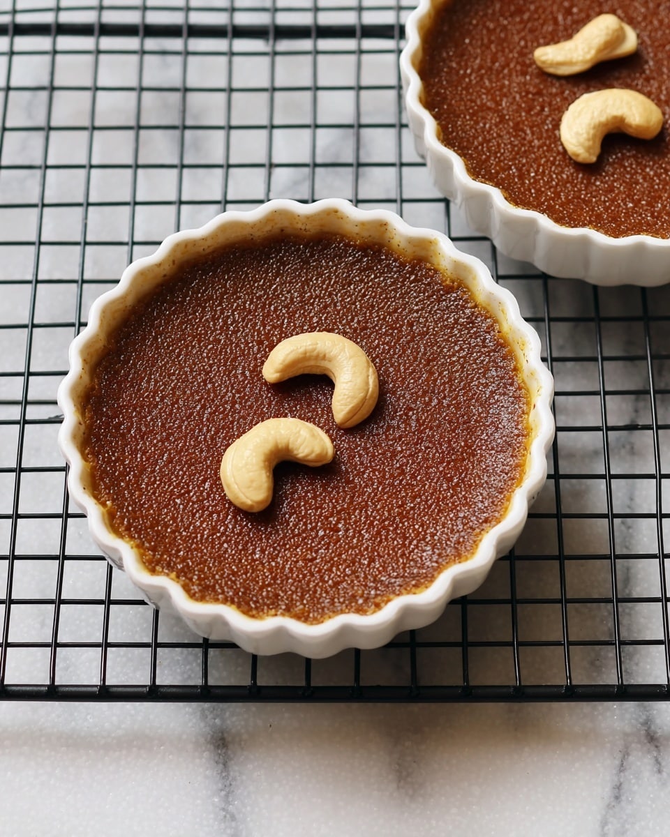 Two small round dishes with scalloped edges, each filled with a smooth, dark brown custard layer that has a slightly grainy texture on top. The custard is creamy and even, topped with three light beige cashew nuts arranged gently in the center of each dish. The dishes are white in color and sit on a black wire cooling rack, which rests on a white marbled surface. The scene is simple and clean, focusing on the rich color contrast between the custard, the cashews, and the white dishes. Photo taken with an iphone --ar 4:5 --v 7