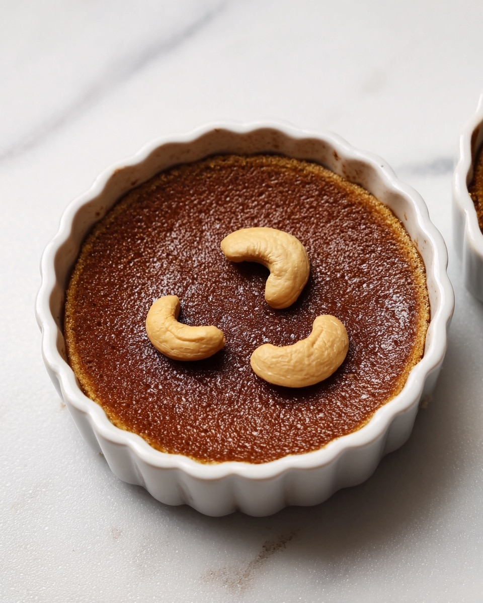 The image shows a single round dessert in a white ceramic fluted dish with a smooth, dark brown top layer that looks glossy and slightly textured. On top, there are four whole cashew nuts placed near the center in a loose circle. The dessert sits on a round surface with a worn look, but the surrounding background is changed to a white marbled texture. The dessert appears to be one layer with the cashews adding a bit of height. photo taken with an iphone --ar 4:5 --v 7